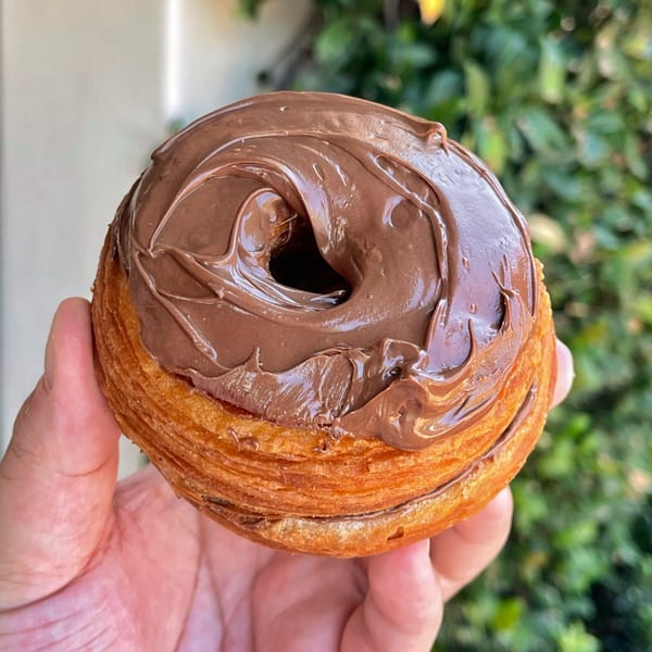 A hand holds a cronut from Mag's Donuts & Bakery, topped with rich chocolate frosting.