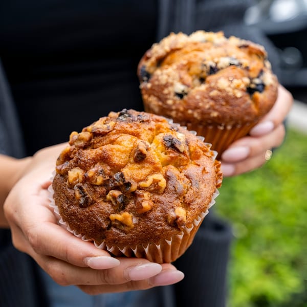 Hands holding two freshly baked muffins, one with walnuts and the other with blueberries and streusel.
