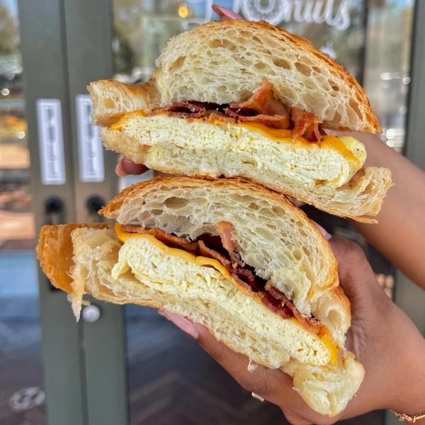 Hands holding a croissant breakfast sandwich with egg, cheese, and bacon, split in half.