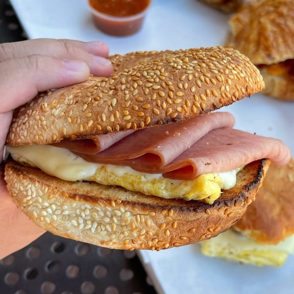 Hand holding a sesame seed bagel sandwich with ham, melted cheese, and scrambled egg.