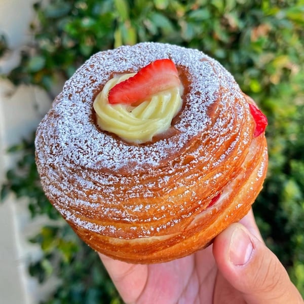 Hand holding a cronut from Mag's Donuts & Bakery, topped with cream, strawberry, and powdered sugar.