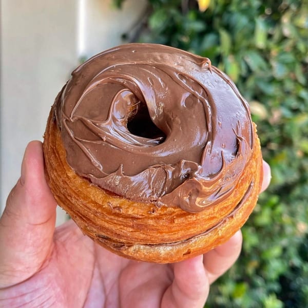Hand holding a cronut from Mag's Donuts & Bakery, topped with rich chocolate frosting.
