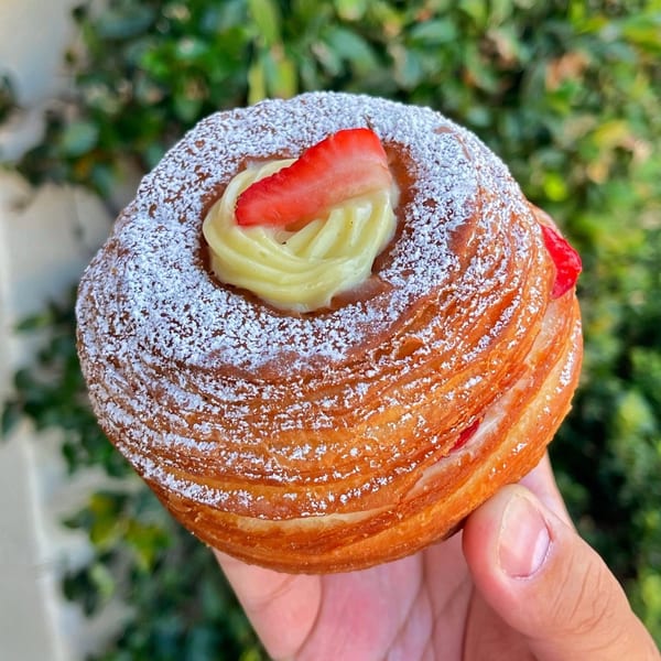 A hand holds a cronut from Mag's Donuts & Bakery, topped with cream, strawberry, and powdered sugar.