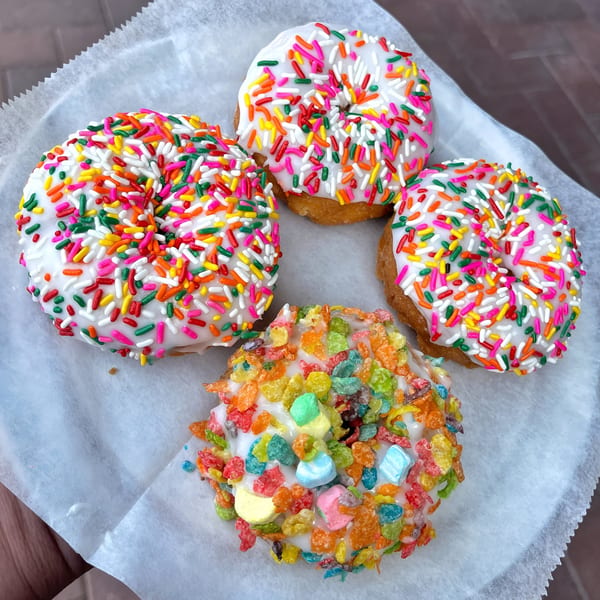 Four Mag's Donuts, three with colorful sprinkles and one with Fruity Pebbles and marshmallows, on white paper.