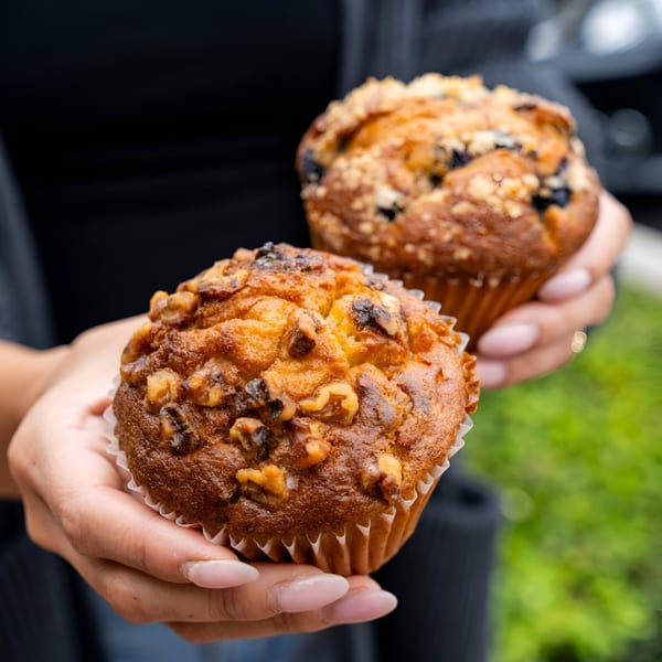 Hands holding two freshly baked muffins, one walnut and one blueberry, from Mag's Donuts & Bakery.