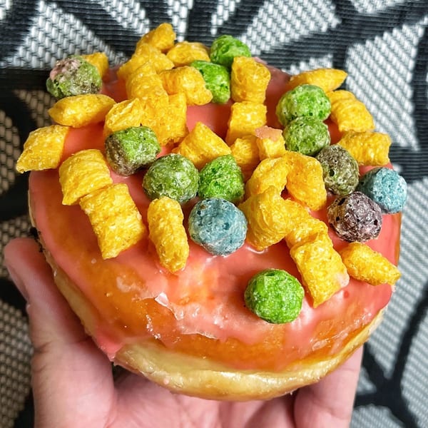 A hand holds a Mag's Donuts & Bakery donut with pink icing and colorful cereal toppings.