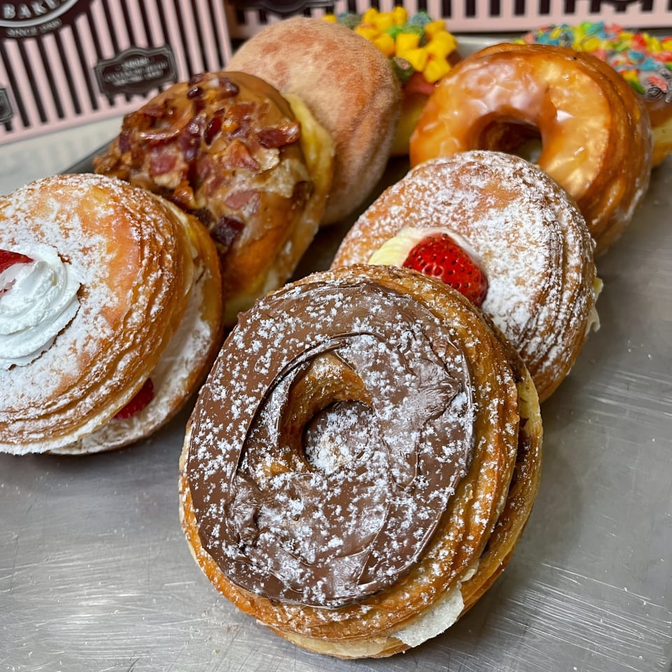 A close-up of various Mag's Donuts & Bakery cronuts, including chocolate, strawberry, and glazed.