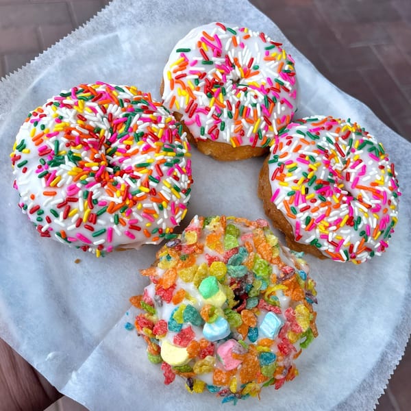 Four Mag's Donuts, three with white icing and rainbow sprinkles, one with Fruity Pebbles cereal.