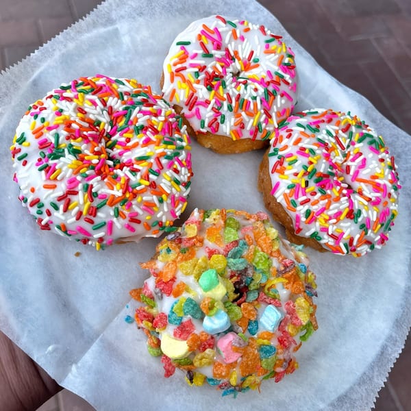 Four Mag's Donuts, three with white frosting and rainbow sprinkles, one with Fruity Pebbles cereal.