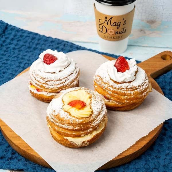 Three Mag's Donuts cronuts with cream, strawberries, and powdered sugar on a wooden board.