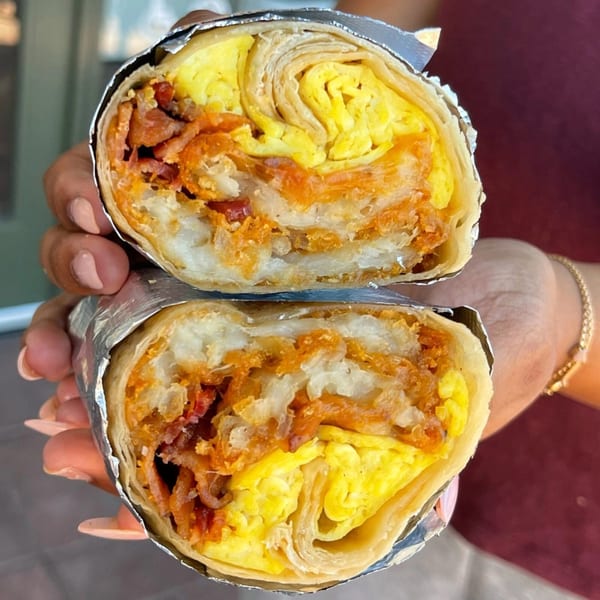 Hands holding a Mag's Donuts & Bakery breakfast burrito, cut in half, revealing eggs, bacon, and hash browns.